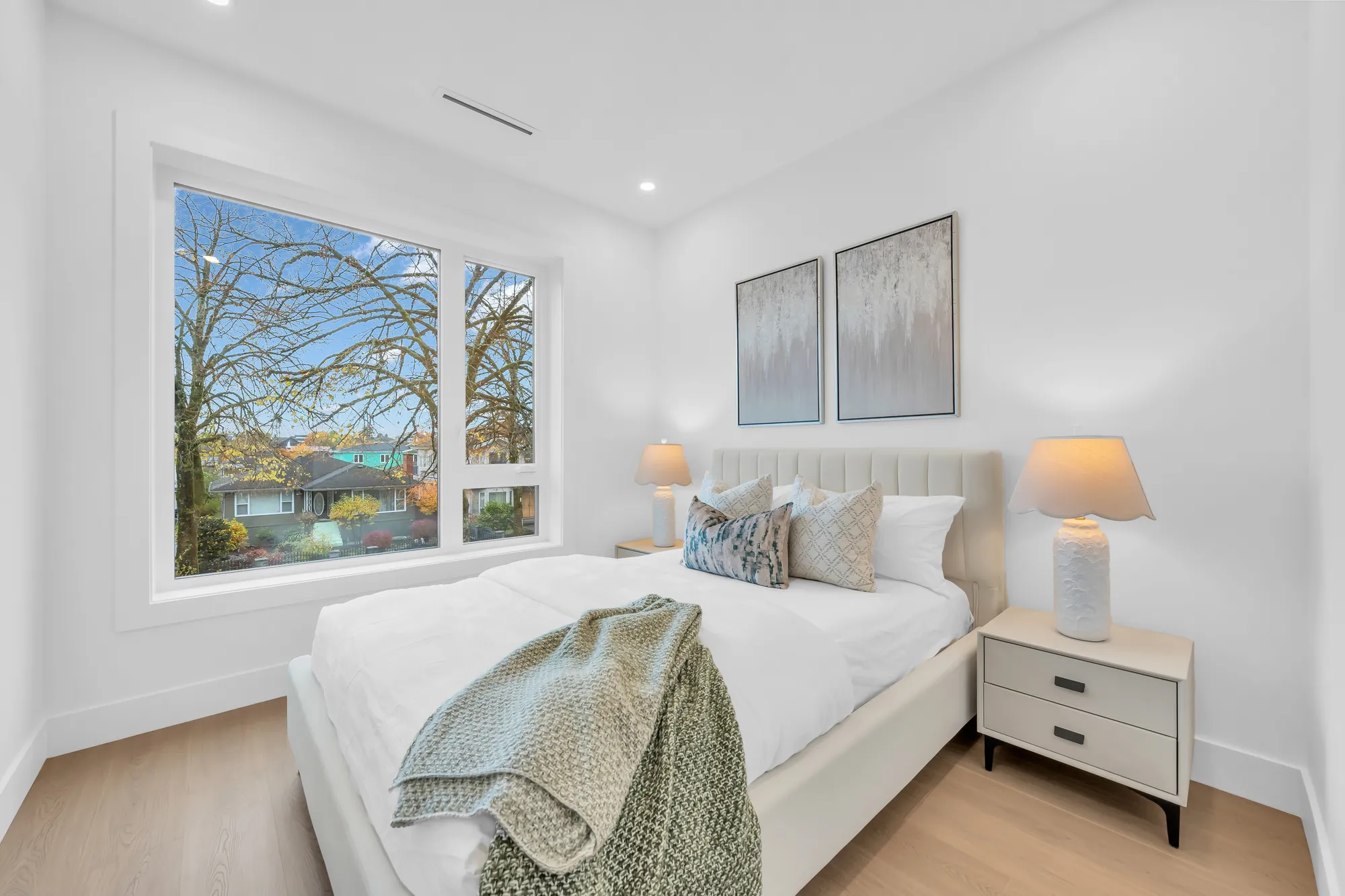 A bedroom: a beige upholstered bed with a green throw, night tables and lamps, two framed prints, a large window onto the trees