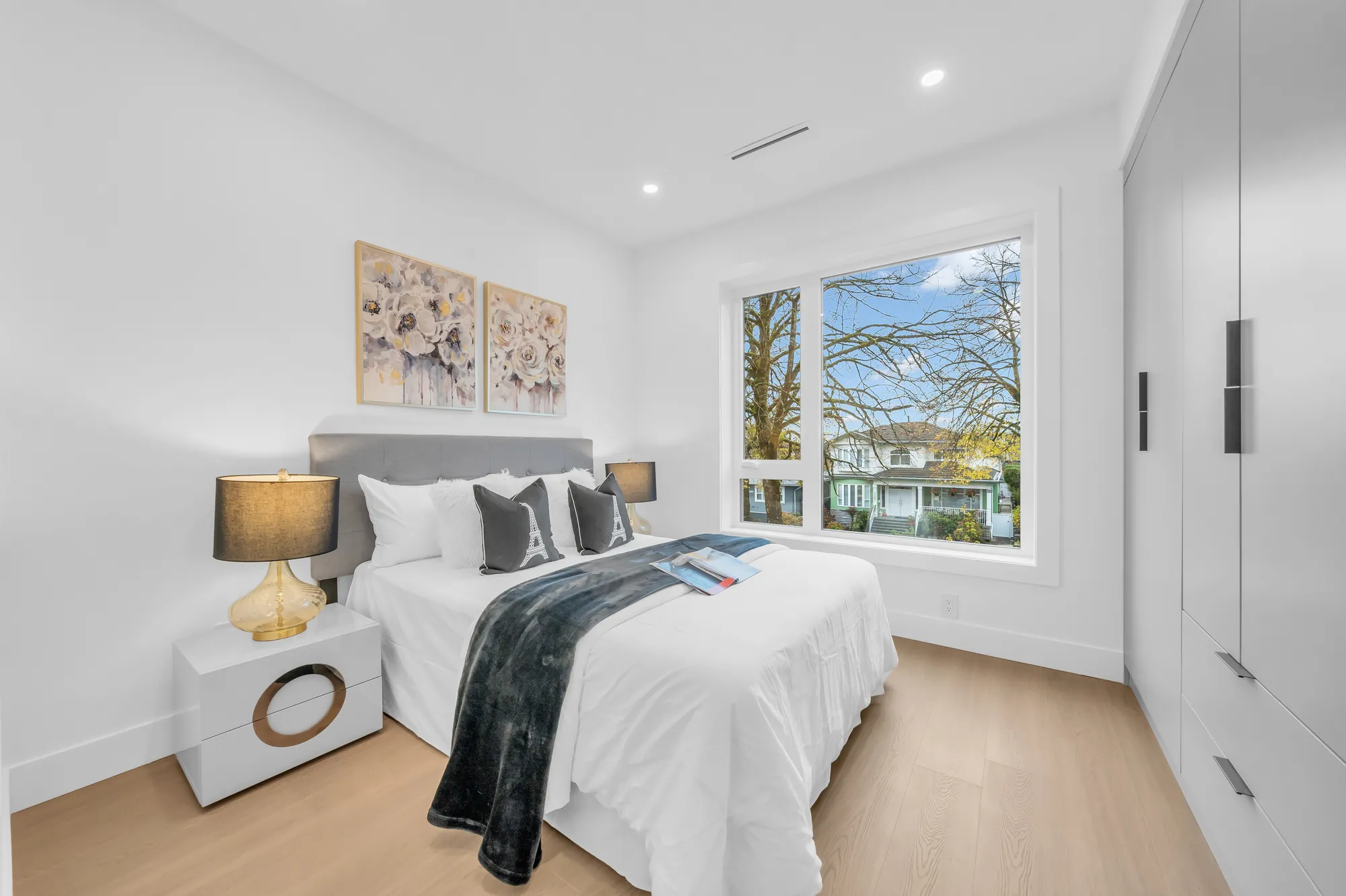 A bedroom: a grey upholstered bed with navy and white bedding, white night tables and lamps, a built-in closet, a window