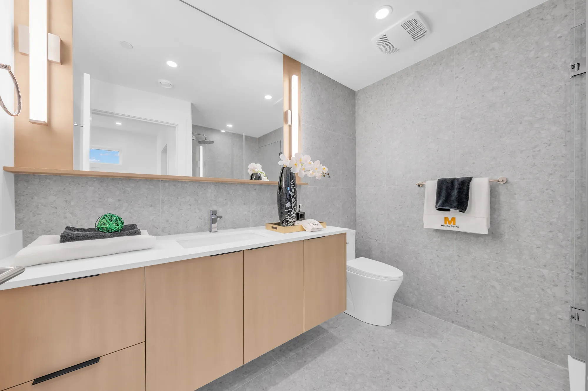 The primary bathroom: a long oak vanity with a white top, a wide mirror and a tall light, grey terrazzo-look tile, a toilet