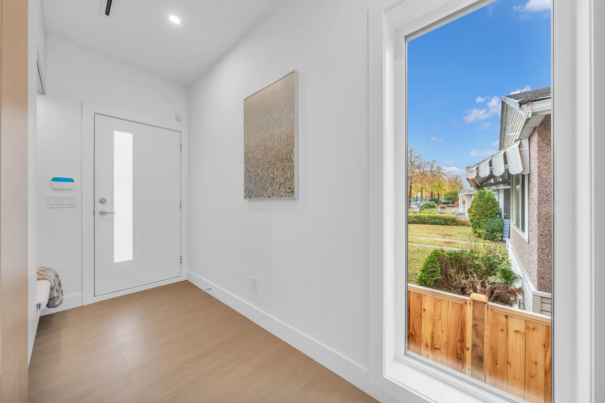 The foyer: a white front door with a slim glass light, a tall window beside it onto the side yard and a wood fence, a pale oak floor