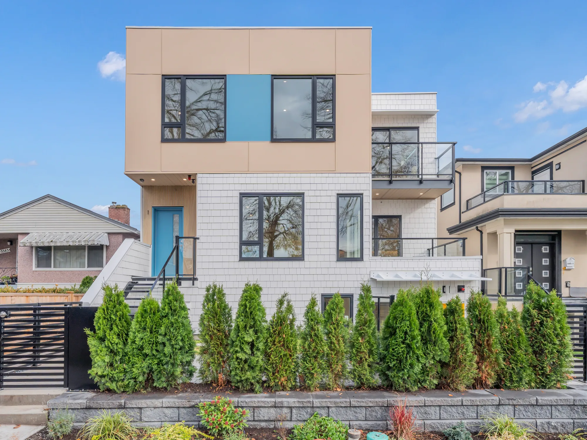 The front of 1-3391 Price Street: a new building with a tan upper floor and white shingle siding, a blue-green panel between two windows and a blue-green door at the top of black steel steps, behind a hedge of cedars