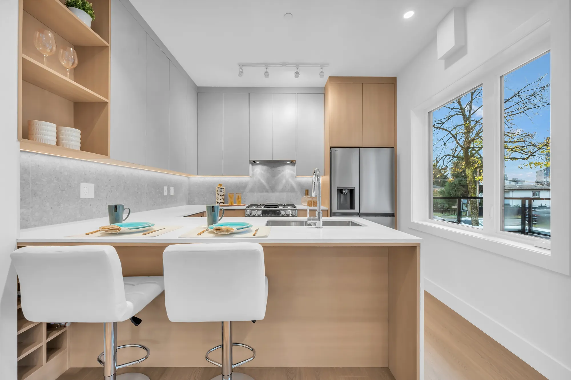 The kitchen: an island with a white waterfall top and two white stools, oak and pale grey cabinets, a stainless fridge, a window at the end