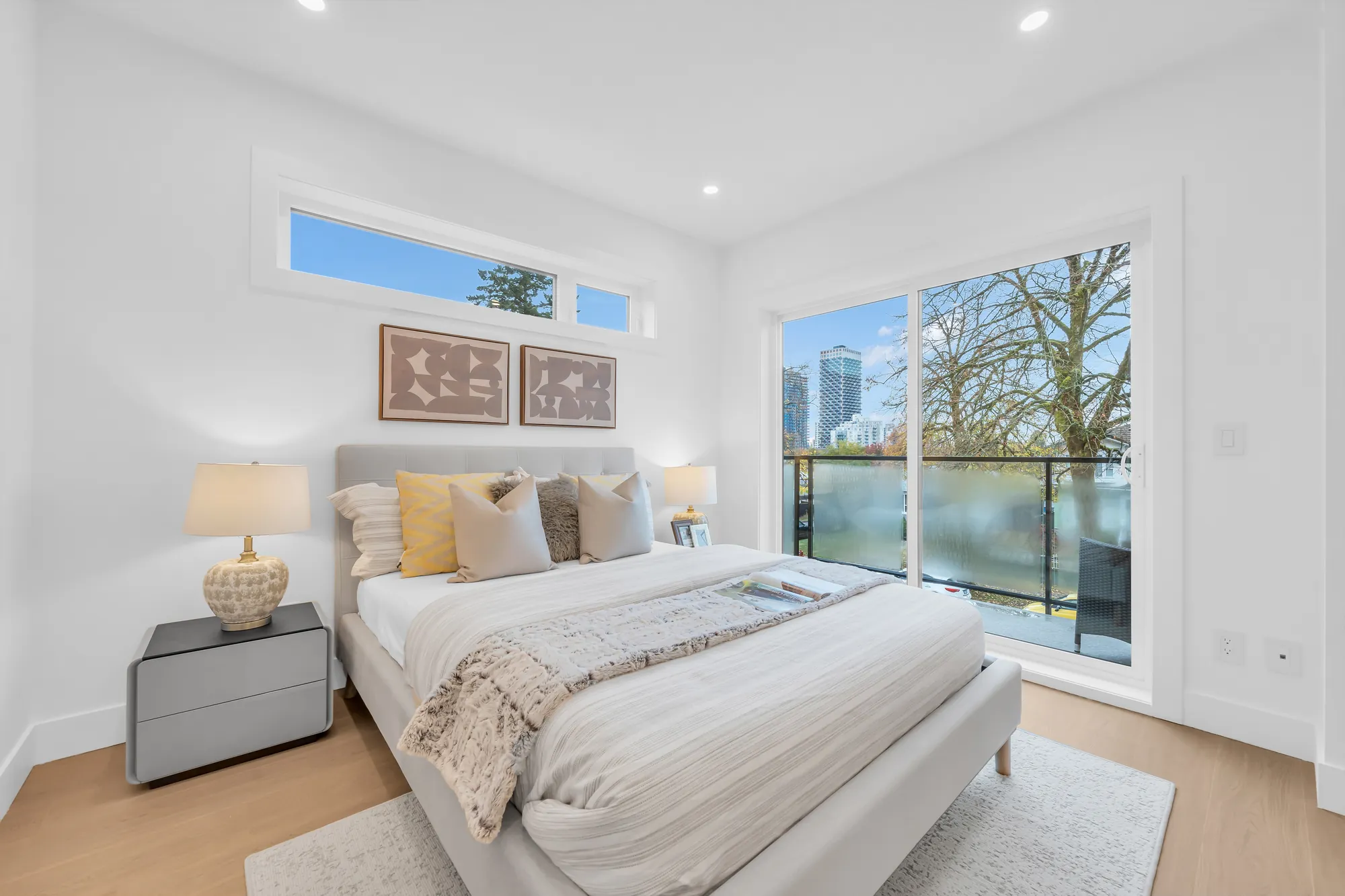 The primary bedroom: a pale upholstered bed with yellow and white cushions, grey night tables and lamps, a clerestory window and a tall glass door to the balcony