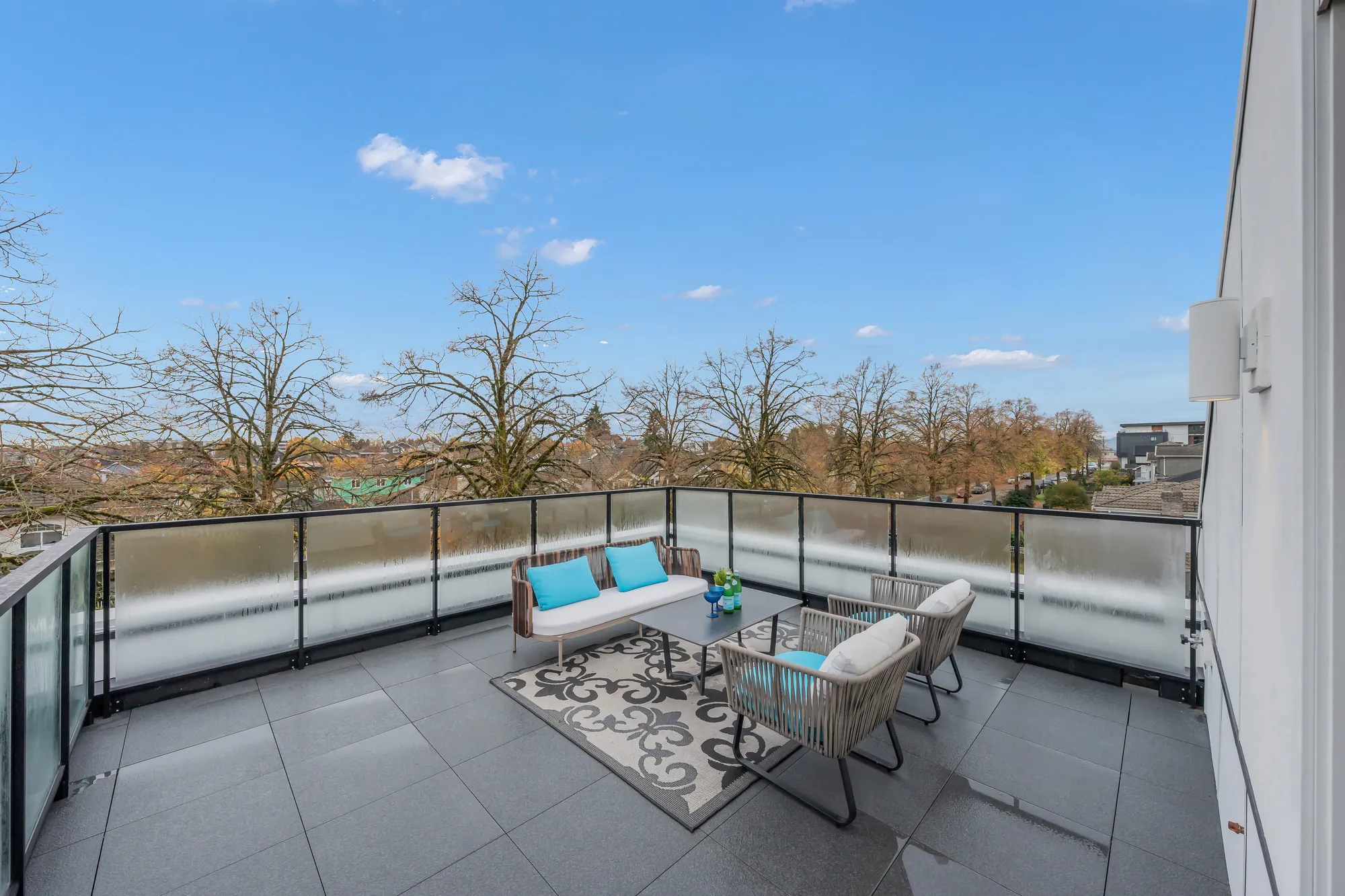 The roof terrace: grey pavers, a glass railing all round, a woven sofa set with blue cushions, treetops and towers beyond